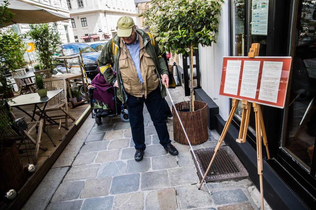 Mann mit Langstock orientiert sich auf dem Gehsteig, Restaurantaufsteller ist eine Barriere