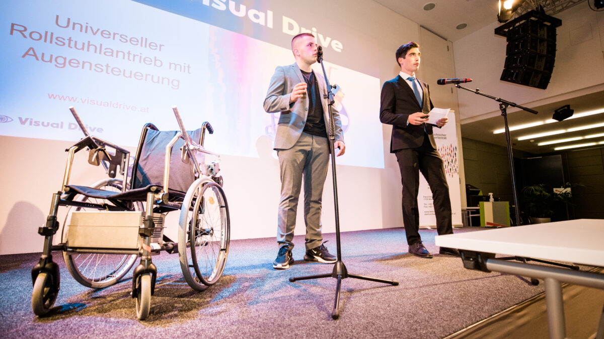 2 Schüler stehen auf der Bühne und präsentieren. Neben ihnen ihr Prototyp, ein adaptierter Rollstuhl.