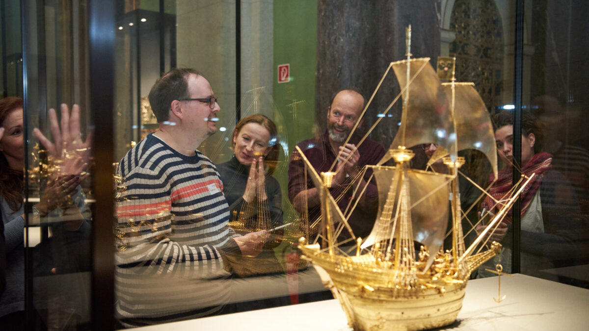 Im Vordergrund sieht man ein goldenes kleines Schiff in einem Glaskasten, dahinter eine Personengruppe die dem Schiff zugewandt ist
