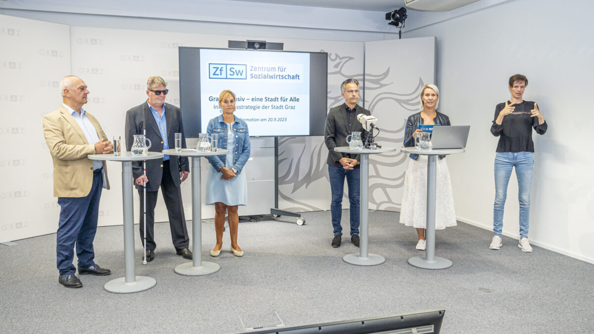 6 Personen stehen vor Stehtischen, eine gebärdet