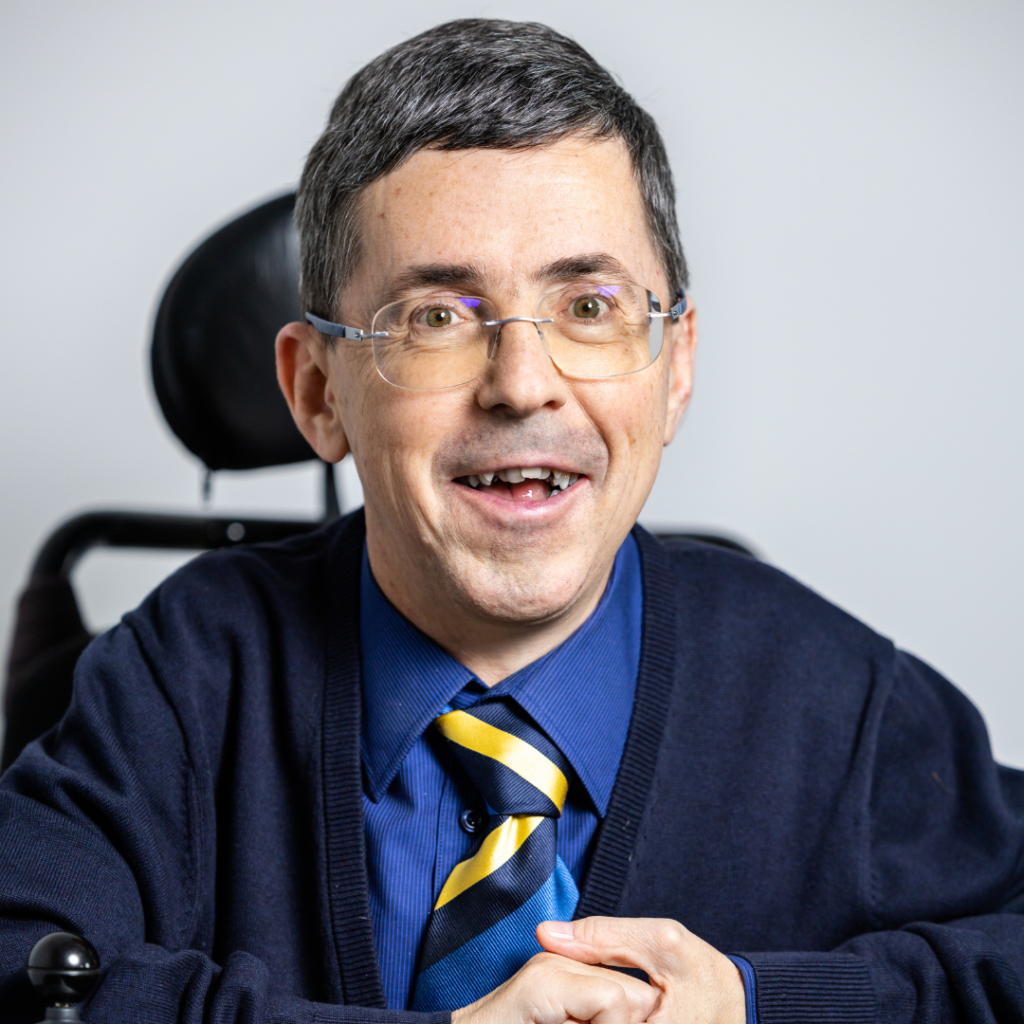 Portrait Martin Ladstätter: Mann mit braun-grauem Haar sitzt in einem E-Rollstuhl, trägt eine blaue Weste und ein blaues Shirt und lächelt freundlich in die Kamera