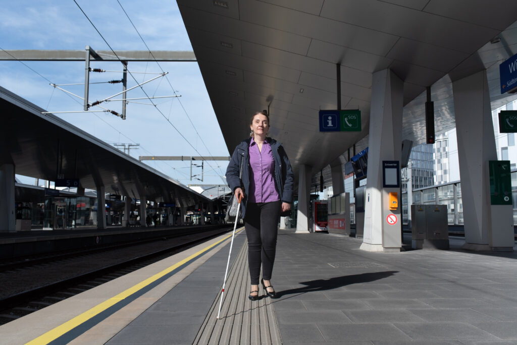 Eine Frau mit weißem Führstock auf einem taktilen Leitsystem am Bahnsteig im ÖBB Hauptbahnhof Wien. Foto: BSVÖ