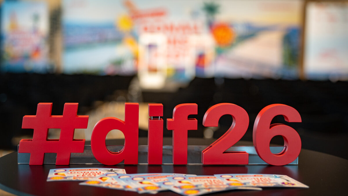 Rotes „#dif26“-Logo auf einem runden Tisch im Vordergrund, dahinter unscharf ein Veranstaltungsbereich mit Bühne, Sitzreihen und Werbebannern des Donauinselfests.