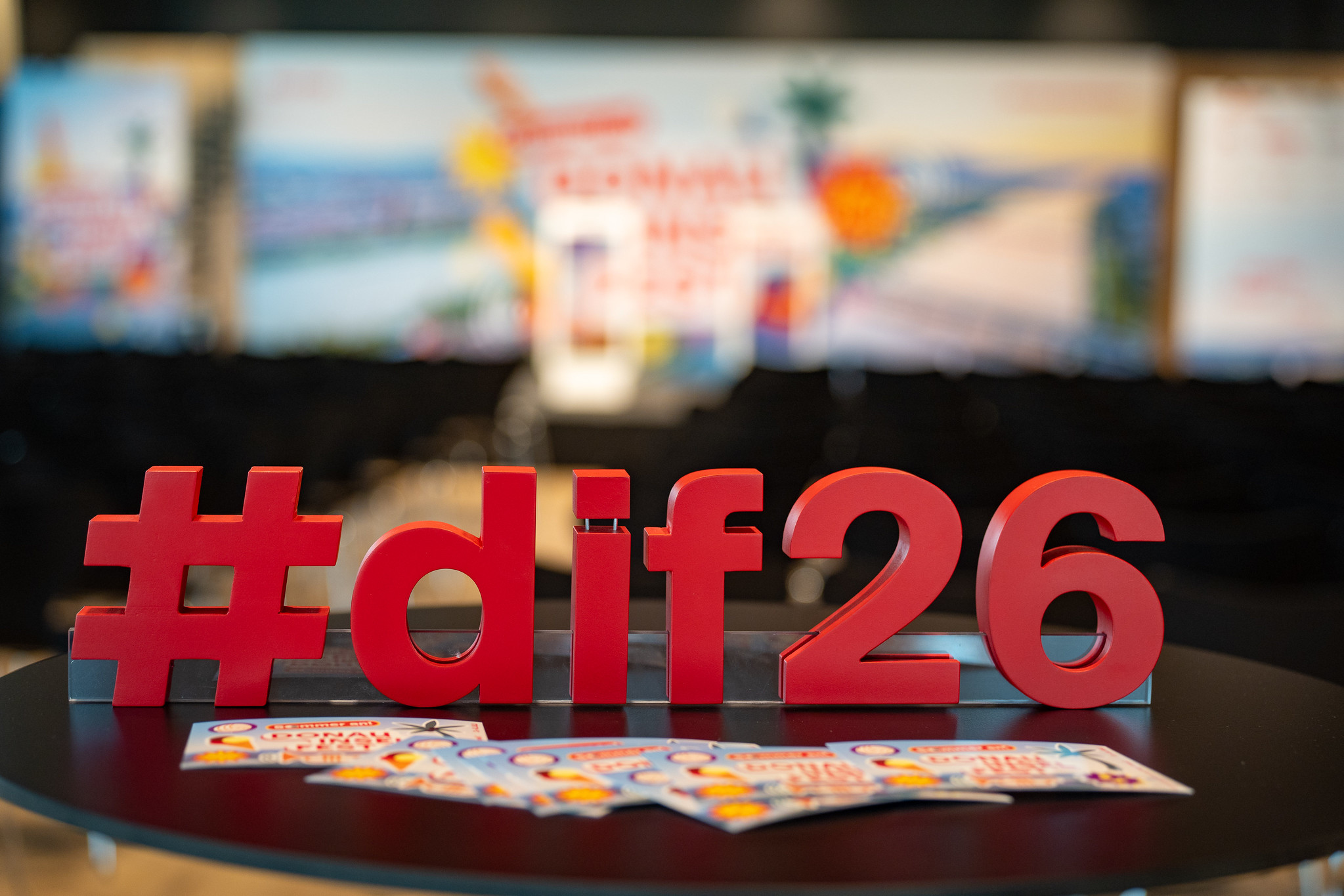 Rotes „#dif26“-Logo auf einem runden Tisch im Vordergrund, dahinter unscharf ein Veranstaltungsbereich mit Bühne, Sitzreihen und Werbebannern des Donauinselfests.