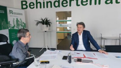 Martin Ladstätter und Helmut Brandstätter unterhalten sich. Sie sitzen an einem Tisch. Im Hintergrund steht an der Wand groß "Behindertenrat" geschrieben.