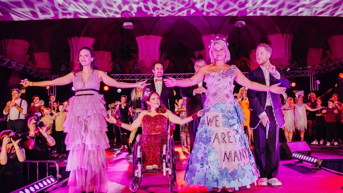 festlich gekleidete Personen mit ausgebreiteten Armen auf einer Bühne beim Diversity Ball
