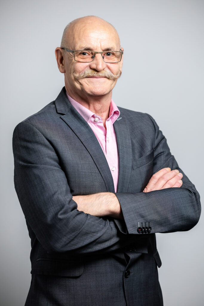 Mann mit brünettem Haar und Schnauzer trägt Brille, rosa Hemd und graues Sakko