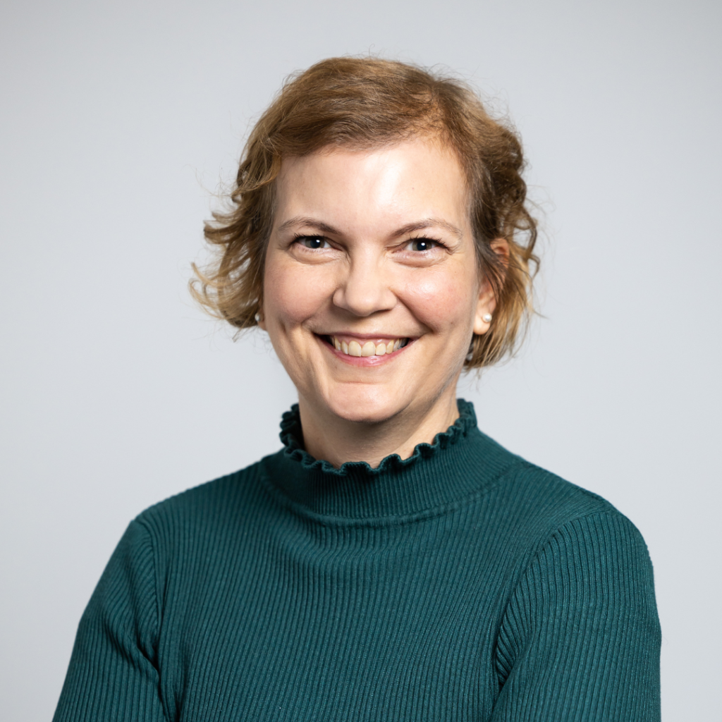 Portraitfoto von Karin Draschkowitz, sie hat rotbraunes, kinnlanges Haar, einen petrollfarbenen Pulli und lächelt in die Kamera