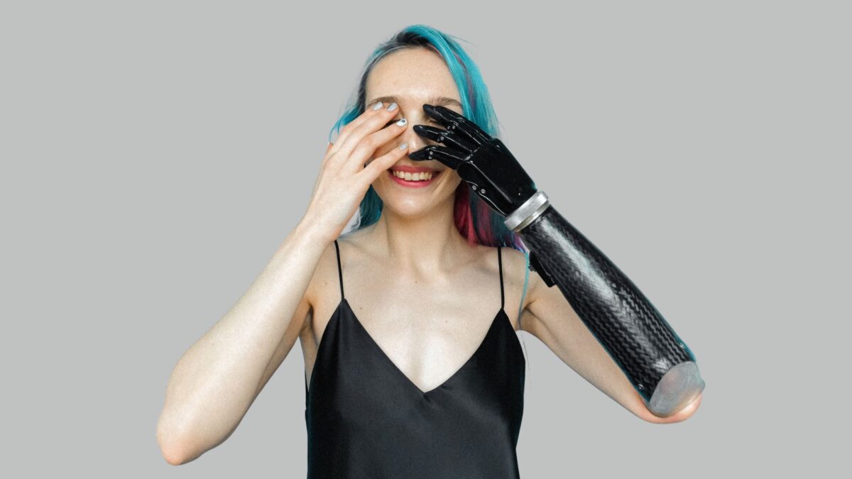 Frau mit blauem Haar und schwarzem Spaghettishirt sowie einer Armprothese einer schwarzen Armprothese mit bionischer Hand verdeckt mit ihren Händen ihre Augen, Foto: Pexels/Anna Shvets
