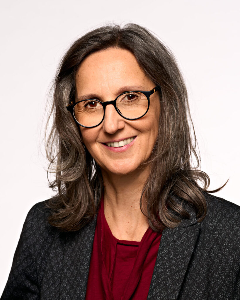 Portrait einer Frau mit braunem, schulterlangem Haar und schwarzer Brille. Sie trägt eine weinrote Bluse und einen grau-melierten Blazer. Sie lächelt in die Kamera