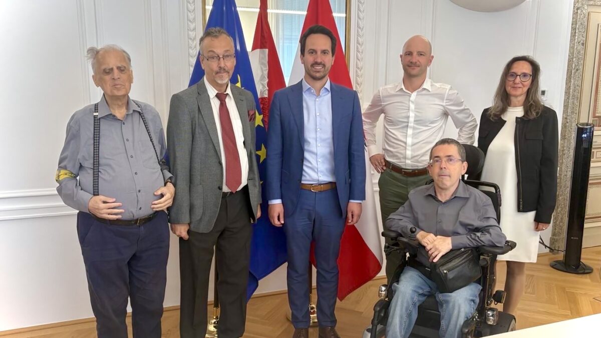 5 stehende und eine im Rollstuhl sitzende Person, der Mann ganz links trägt am rechten Arm eine gelbe Schleife mit drei schwarzen Punkten