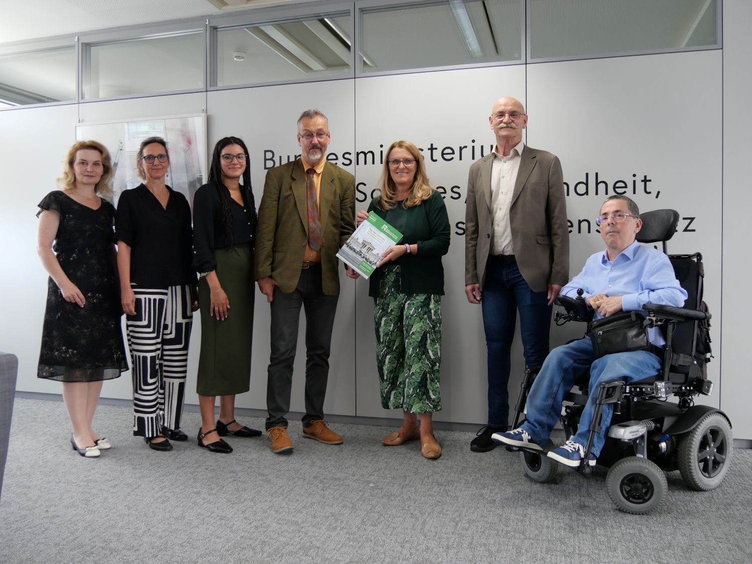 6 stehende Personen und eine im Rollstuhlsitzende Personen vor dem Schild des Sozialministeriums