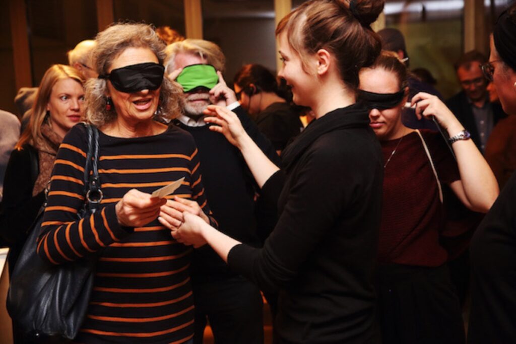 Theaterbesucher*innen mit Dunkelmasken auf den Augen werden von einer Frau ohne Maske geführt