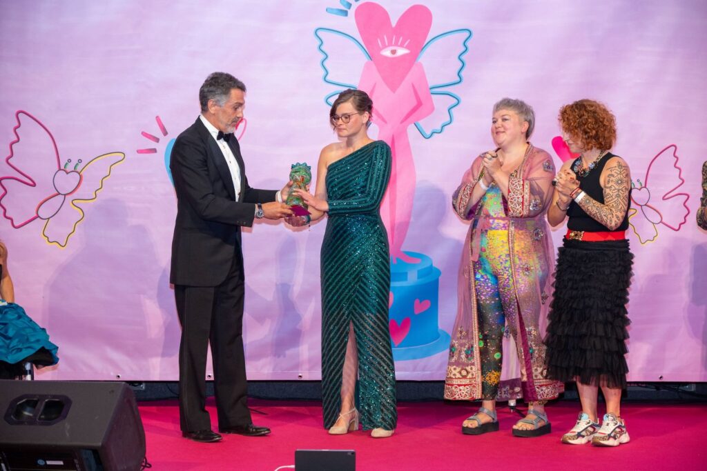 festlich gekleideter Mann übergibt junger Frau mit türkisem Ballkleid eine Statue. Rechts daneben stehen zwei Frauen und klatschen.