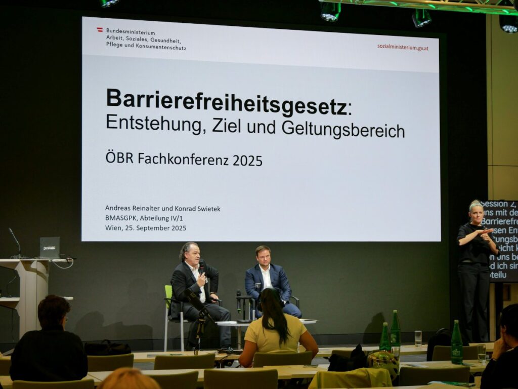 2 Männer sitzen auf Stühlen auf einer Bühne und sprechen vor Publikum, rechts dolmetscht eine ÖGS-Dolmetscherin