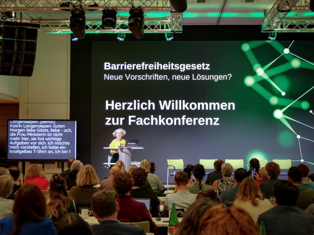 Frau mit grau-braunem Haar, gelber Bluse und grünem Rock steht vor einem Stehpult auf einer Bühne und spricht. Im Vordergrund sitzendes Publikum; Foto: Österreichischer Behindertenrat/Andrea Strohriegl