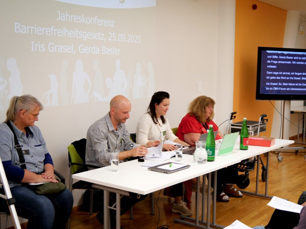 4 vor einem weißen Tisch sitzende Personen, im Hintergrund eine Präsentation "Jahreskonferenz Barrierefreiheitsgesetz, Iris Grasel, Gerda Basler