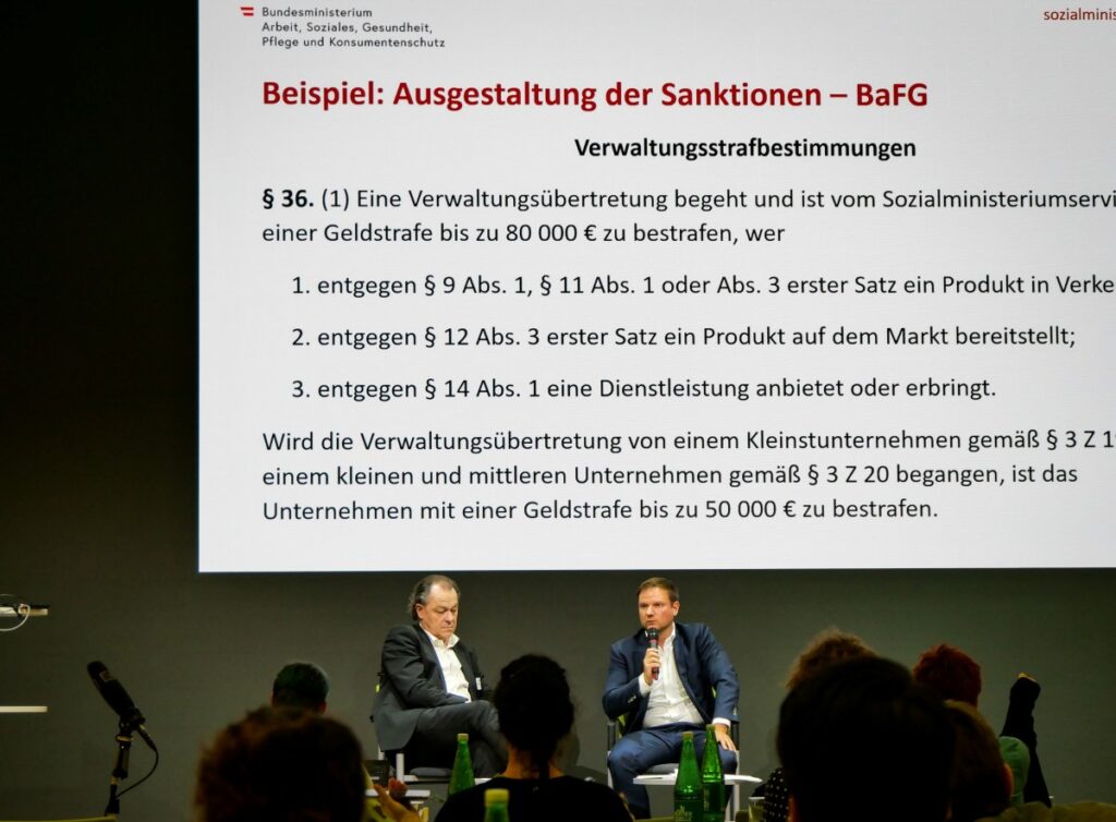 2 Männer sitzen auf Stühlen auf einer Bühne und sprechen vor Publikum. Im Hintergrund Präsentation über "Beispiel: Ausgestaltung Sanktionen - BaFG"