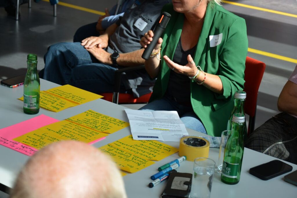 2 Personen, die nicht erkennbar sind, arbeiten mit unterschiedlichen bunten Postit und bunten Zetteln an Thementischen.