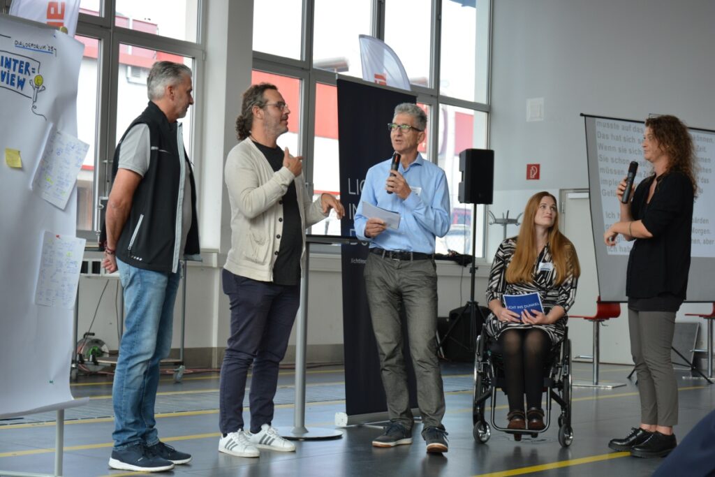 Zwei Mitarbeiter von Rosenbauer International im Gespräch mit den zwei Moderatorinnen beim dritten LICHT INS DUNKEL-Dialogforum zum Thema inklusive Arbeitswelt. Eine ÖGS-Dolmetscherin übersetzt die Wortmeldung einer der beiden Mitarbeiter. Im Hintergrund sieht man die Live-Schriftdolmetschung.