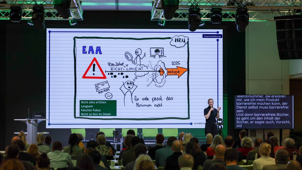 Personen sitzen im Publikum einer Konferenz. Auf einer großen Leinwand ist ein Graphic Recording von Petra Plicka zum European Accessibility Act zu sehen. Rechts auf der Bühne steht eine ÖGS-Dolmetscherin vor einem Bildschirm mit Schriftdolmetschung.