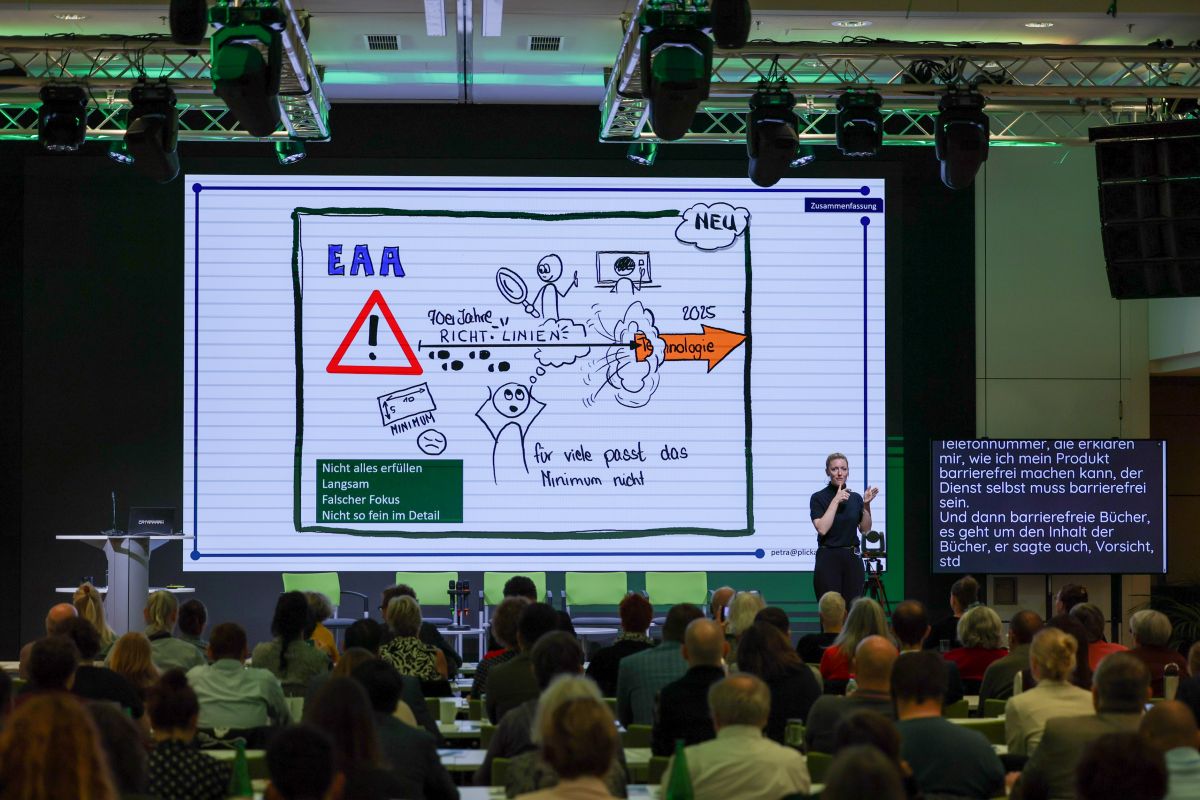 Personen sitzen im Publikum einer Konferenz. Auf einer großen Leinwand ist ein Graphic Recording von Petra Plicka zum European Accessibility Act zu sehen. Rechts auf der Bühne steht eine ÖGS-Dolmetscherin vor einem Bildschirm mit Schriftdolmetschung.