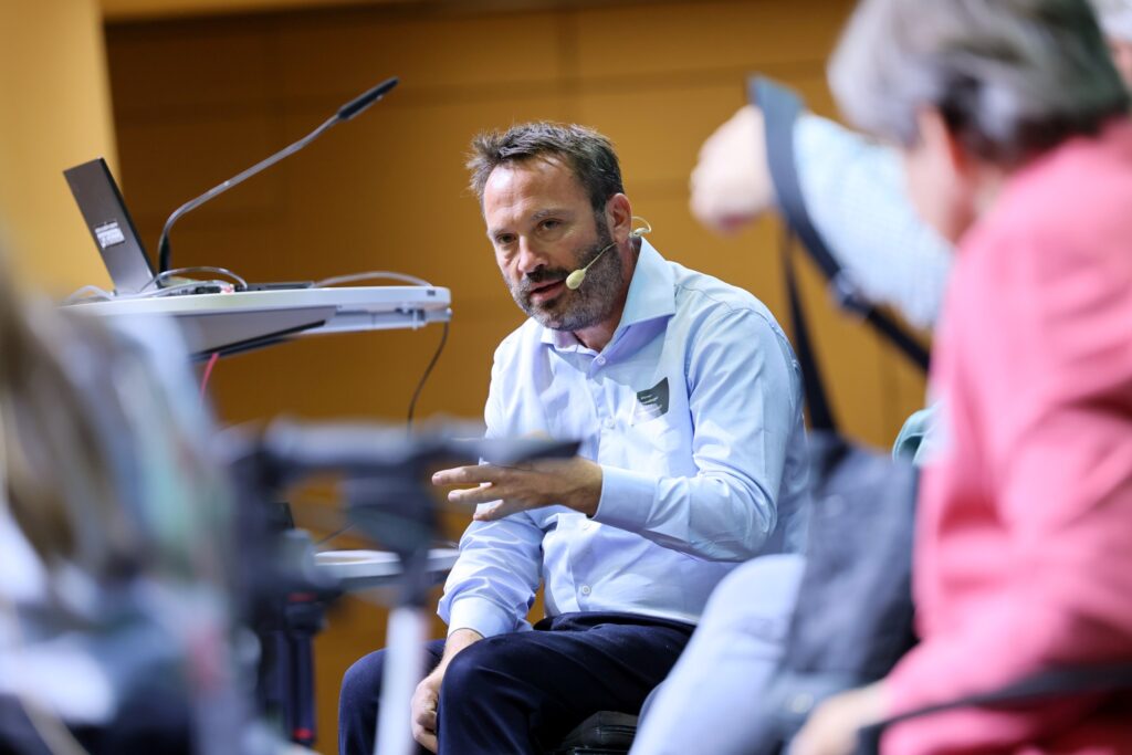 Mann mit braunem Haar und braunem Bart sitzt auf einer Bühne in einem Rollstuhl. Er trägt Jeans und ein blaues Hemd und spricht in ein Headset-Mikrofon