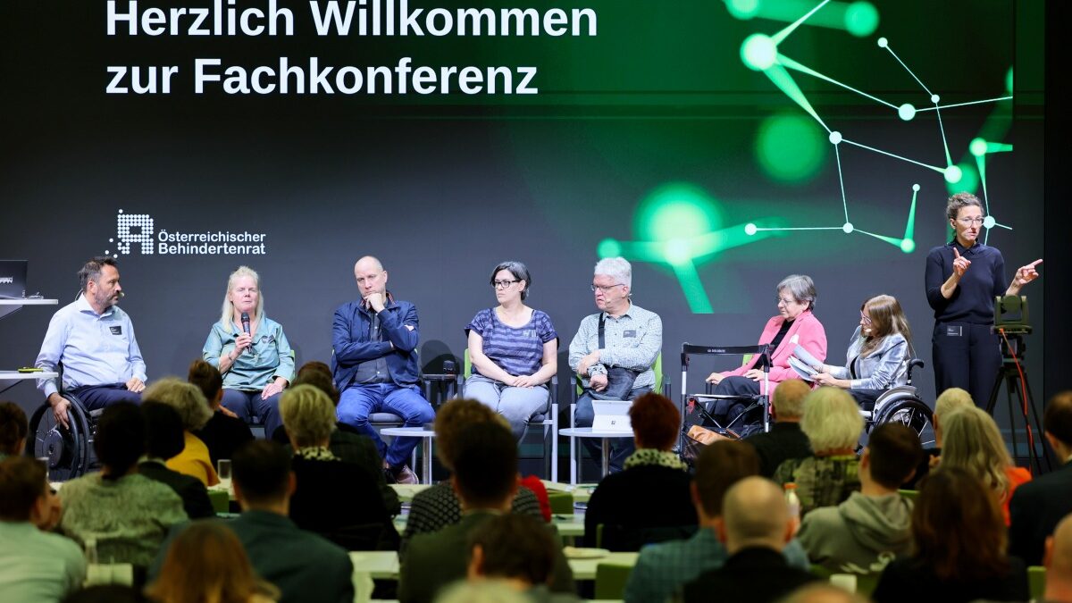 7 sitzende Personen bei einer Podiumsdiskussion. Recht steht eine schwarz gekleidete ÖGS-Dolmetscherin. Im Vordergrund ist sitzendes Publikum zu sehen. Im Hintergrund großer Schriftzug "Herzlich willkommen zur Fachkonferenz"