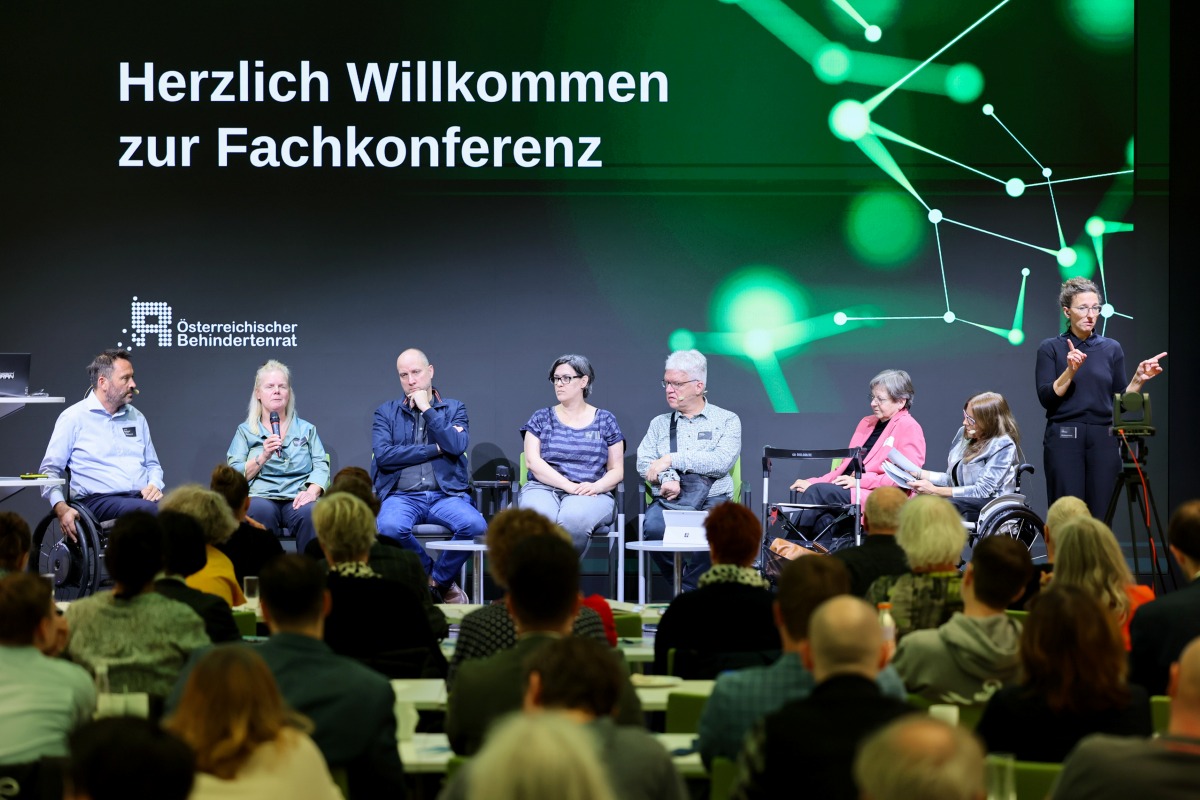 7 sitzende Personen bei einer Podiumsdiskussion. Recht steht eine schwarz gekleidete ÖGS-Dolmetscherin. Im Vordergrund ist sitzendes Publikum zu sehen. Im Hintergrund großer Schriftzug "Herzlich willkommen zur Fachkonferenz"