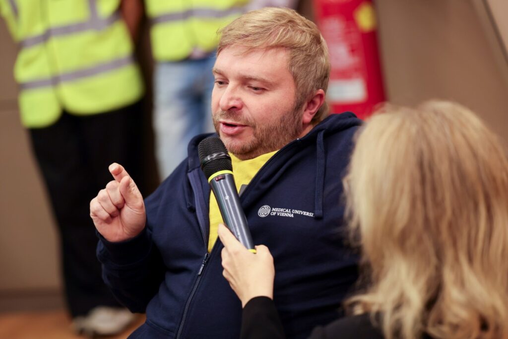 Mann mit blondem Haar und blondem Bart, blauer Weste und gelbem Shirt spricht in ein Mikrofon