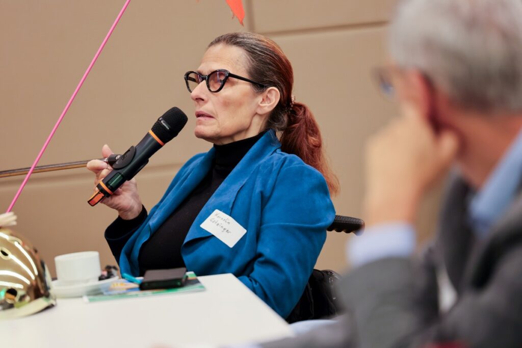 Frau mit langem, rot-braunem Haar, schwarzer Brille, schwarzem Pullover und blauem Blazer spricht sitzend in ein Mikrofon
