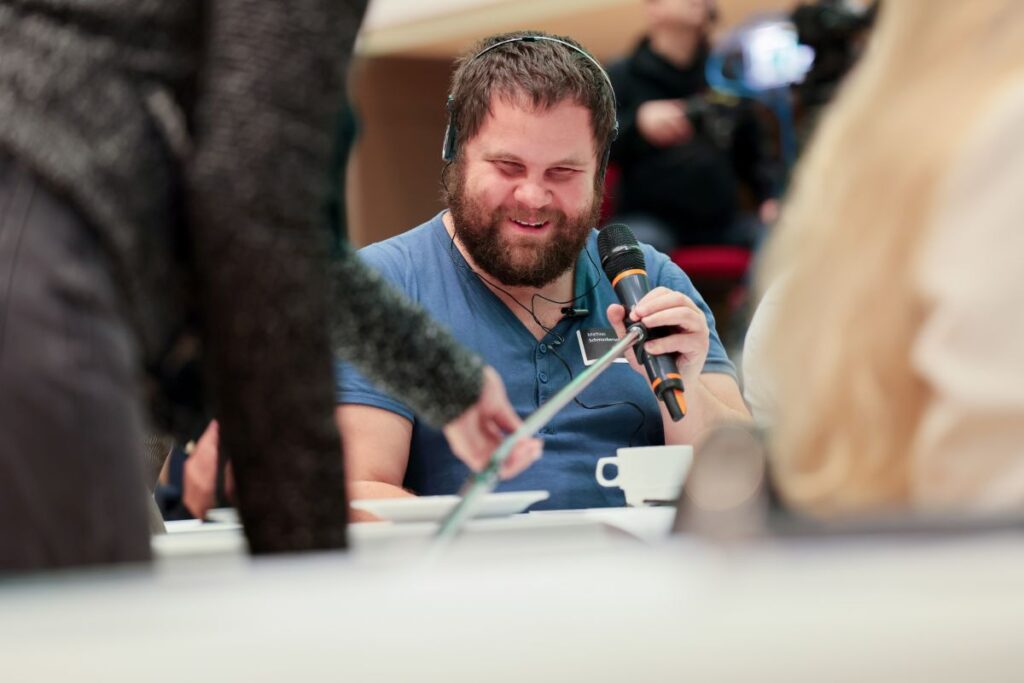 Mann mit braunem Haar, braunem Vollbart und blauem Shirt sitzt in einem Veranstaltungsraum und spricht in ein Mikrofon
