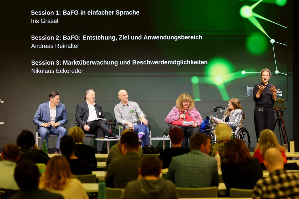 5 sitzende Personen auf einer Bühne, daneben eine stehende, schwarz gekleidet ÖGS-Dolmetscherin. Text im Hintergrund: „Session 1: BaFG in einfacher Sprache. Iris Grasel. Session 2: BaFG: Entstehung, Ziel und Anwendungsbereich. Andreas Reinalter. Session 3: Marktüberwachung und Beschwerdemöglichkeiten. Nikolaus Eckereder“
