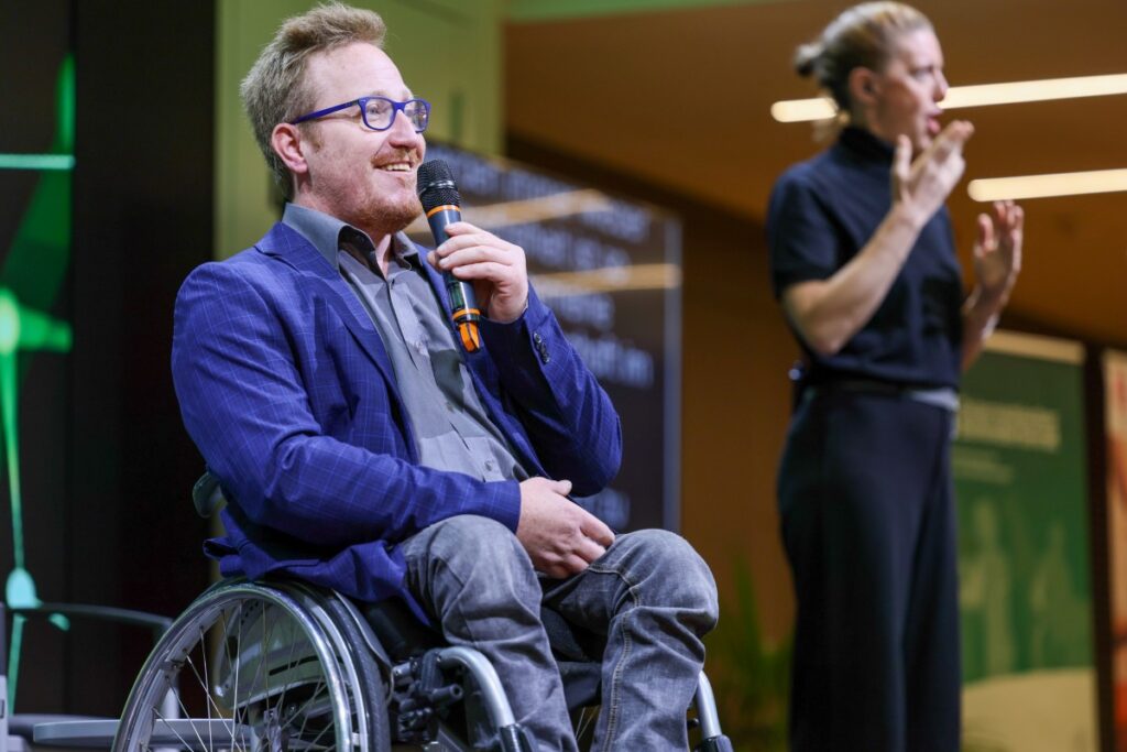 Mann mit blondem Haar, rotem Bart, blauer Brille, blauem Sakko, grauem Hemd und grauer Hose sitzt in einem Rollstuhl und spricht in ein Mikrofon