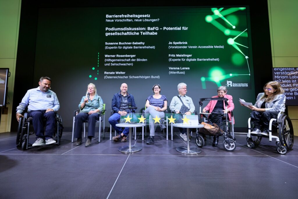 8 Personen sitzen auf einer Bühne. 2 der Personen nützen einen Rollstuhl, eine einen Rollator