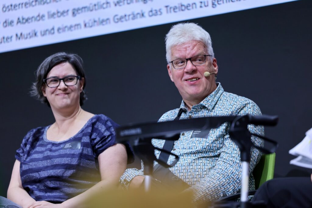 Frau mit braunem, kinnlangem Haar, schwarzer Brille und blau-grau-gestreiftem Shirt sitzt neben einem Mann mit grauem Haar, schwarzer Brille und bunt gemustertem Hemd