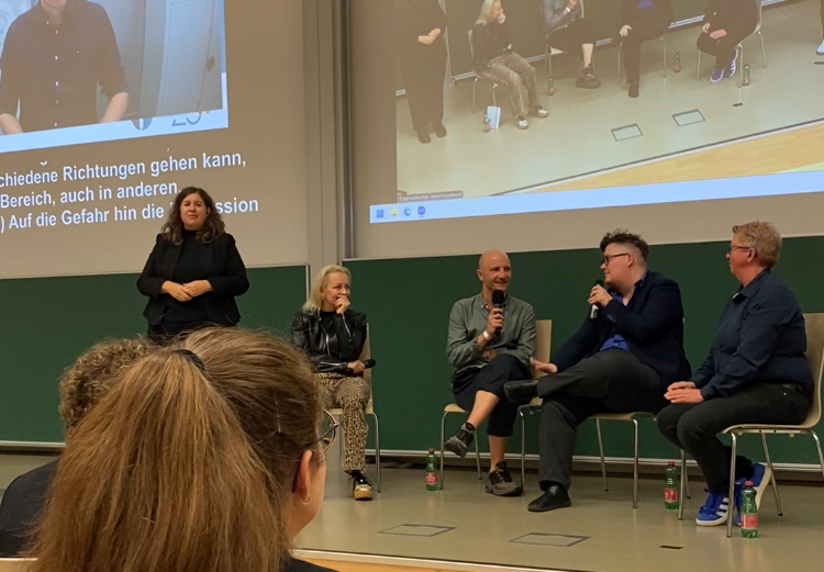 Podium bei einem Kongress mit mehreren Personen auf der Bühne. Die Podiumsteilnehmer*innen sitzen und sprechen. Links eine schwarz gekleidete Gebärdensprach-Dolmetscher*in