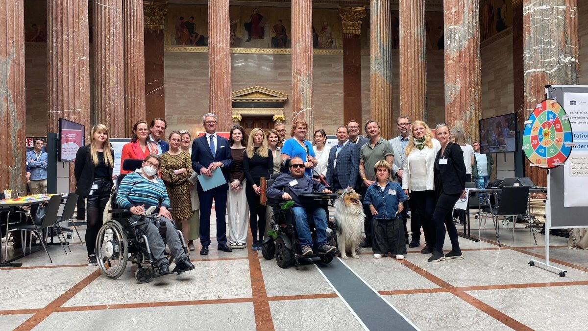 Gruppenfoto bei den Sensibilisierungstagen Barrierefreiheit in der Säulenhalle des Parlaments. Mehrere Personen stehen gemeinsam zwischen hohen Marmorsäulen, darunter Menschen mit und ohne Behinderungen. Zwei Personen nutzen Rollstühle, außerdem ist ein Assistenzhund zu sehen. Rund um die Gruppe befinden sich Informationsstände und Bildschirme.