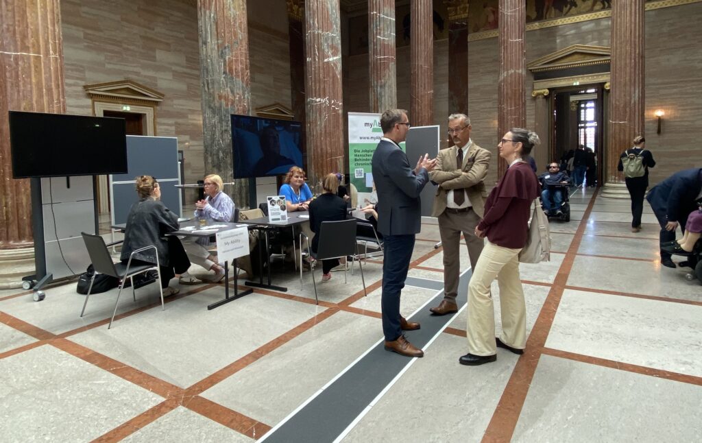 Große Halle im Parlament mit hohen Marmorsäulen während der Sensibilisierungstage Barrierefreiheit. Mehrere Personen stehen oder sitzen an Informationsständen im Gespräch. Im Hintergrund bewegt sich eine Person mit Elektrorollstuhl durch die Halle.