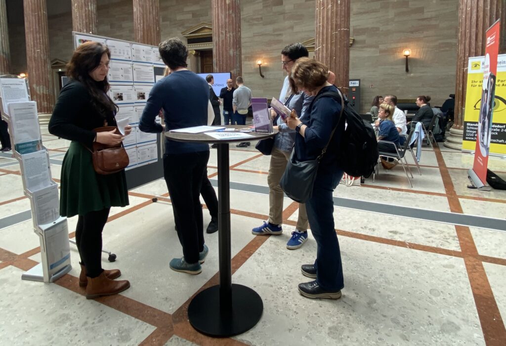 Besucherinnen informieren sich bei den Sensibilisierungstagen Barrierefreiheit in der Säulenhalle des Parlaments an Informationsständen und Schautafeln. Mehrere Personen lesen Unterlagen oder führen Gespräche. Im Hintergrund sitzen weitere Besucherinnen an Tischen zwischen den hohen Marmorsäulen der Halle.