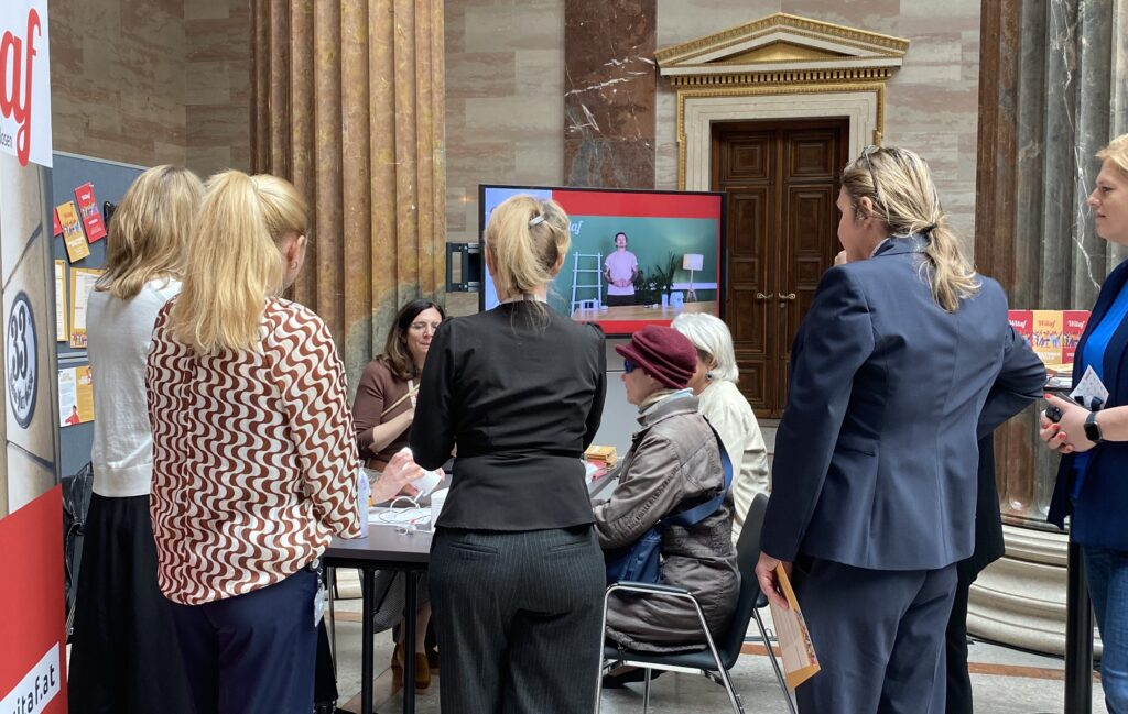 Besucher*innen informieren sich am Stand von WITAF bei den Sensibilisierungstagen Barrierefreiheit in der Säulenhalle des Parlaments. Mehrere Personen stehen und sitzen im Gespräch rund um einen Informationstisch. Im Hintergrund ist ein Bildschirm mit Präsentation zu sehen. Eine blinde Person mit dunkler Brille sitzt am Tisch.
