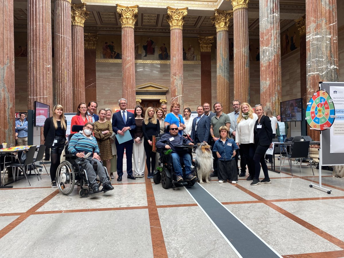 Gruppenfoto bei den Sensibilisierungstagen Barrierefreiheit in der Säulenhalle des Parlaments. Mehrere Personen stehen gemeinsam zwischen hohen Marmorsäulen, darunter Menschen mit und ohne Behinderungen. Zwei Personen nutzen Rollstühle, außerdem ist ein Assistenzhund zu sehen. Rund um die Gruppe befinden sich Informationsstände und Bildschirme.