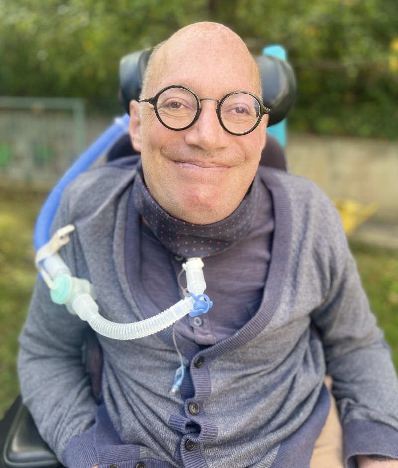 lächelnder Mann mit runder, dunkler Brille, Glazte, bekleidet mit lila Weste und lila Schal sitzt in einem Rollstuhl. Er nützt ein Beatmungsgerät.