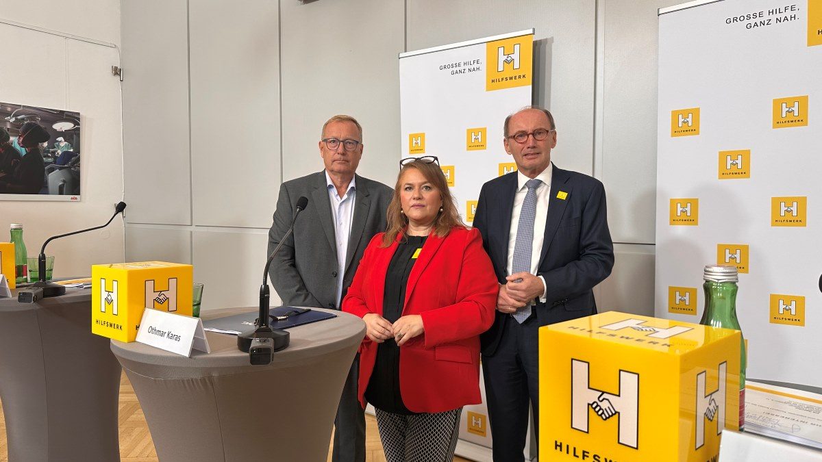 2 Männer mit dunklen Anzügen und eine Frau mit langen, braunen Haaren, schwarz-weißer Hose und rotem Blazer bei einer Pressekonferenz