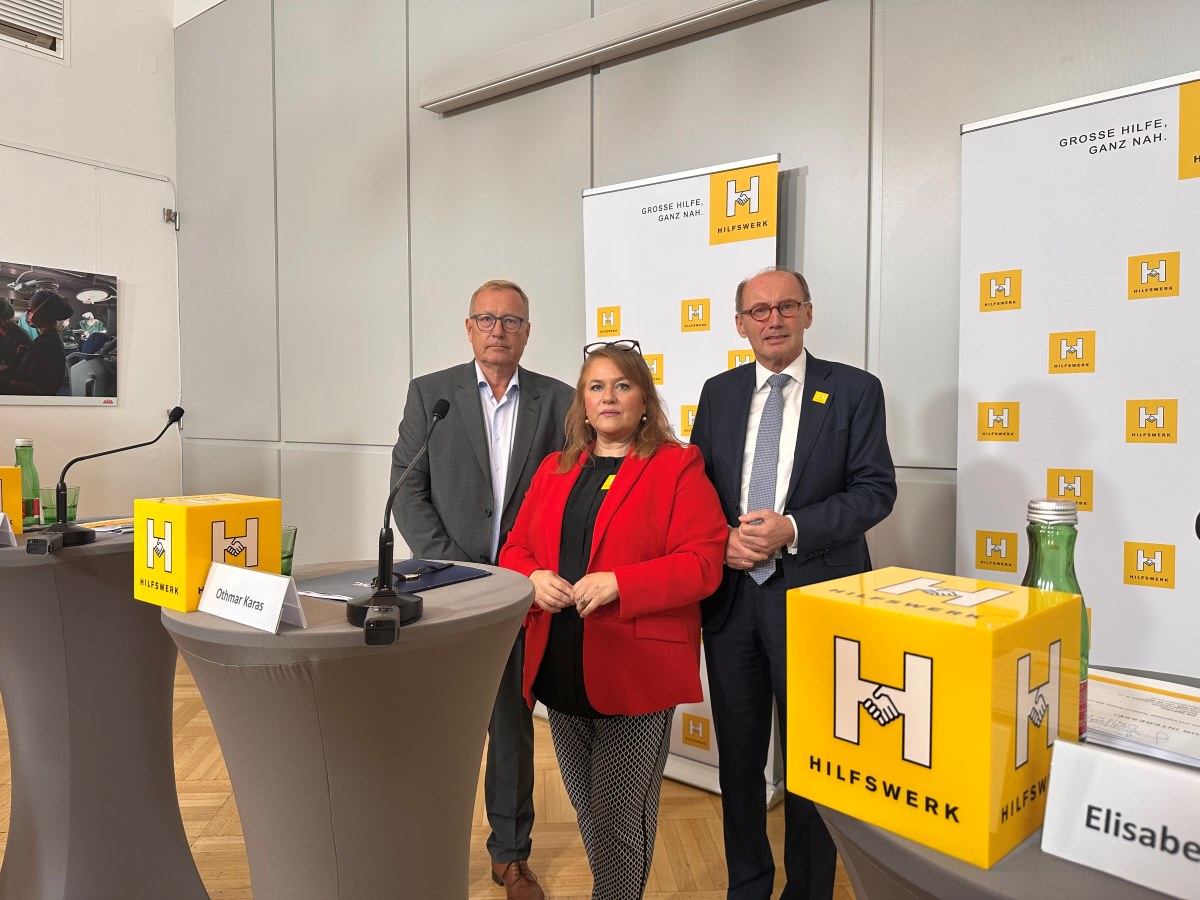 2 Männer mit dunklen Anzügen und eine Frau mit langen, braunen Haaren, schwarz-weißer Hose und rotem Blazer bei einer Pressekonferenz