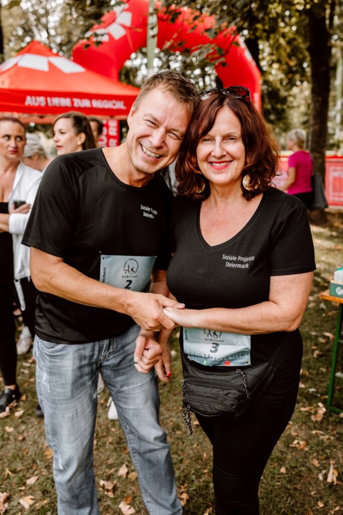ein Mann mit schwarzem Shirt und Jeans und eine Freu mit schwarzem Shirt und schwarzer Hose halten sich bei einem Laufbewerb an den Händen