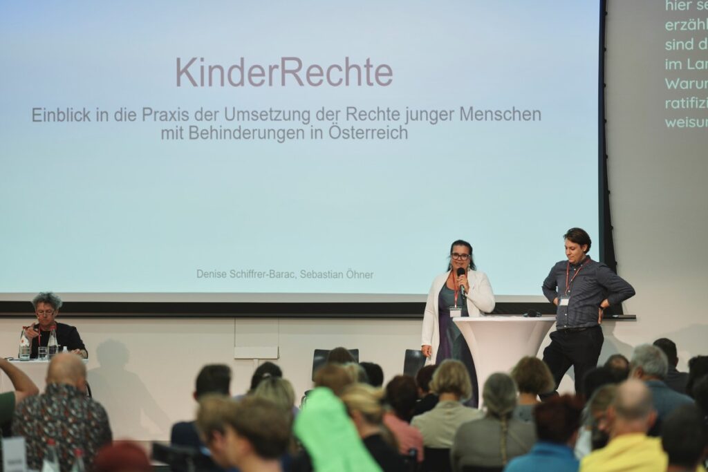 2 Personen auf Bühne sprechen ins Mikro: Mann mit kurzen dunklen Haaren und E-Rolli, Frau mit kinnlangen blonden Haaren und beigem Cardigan