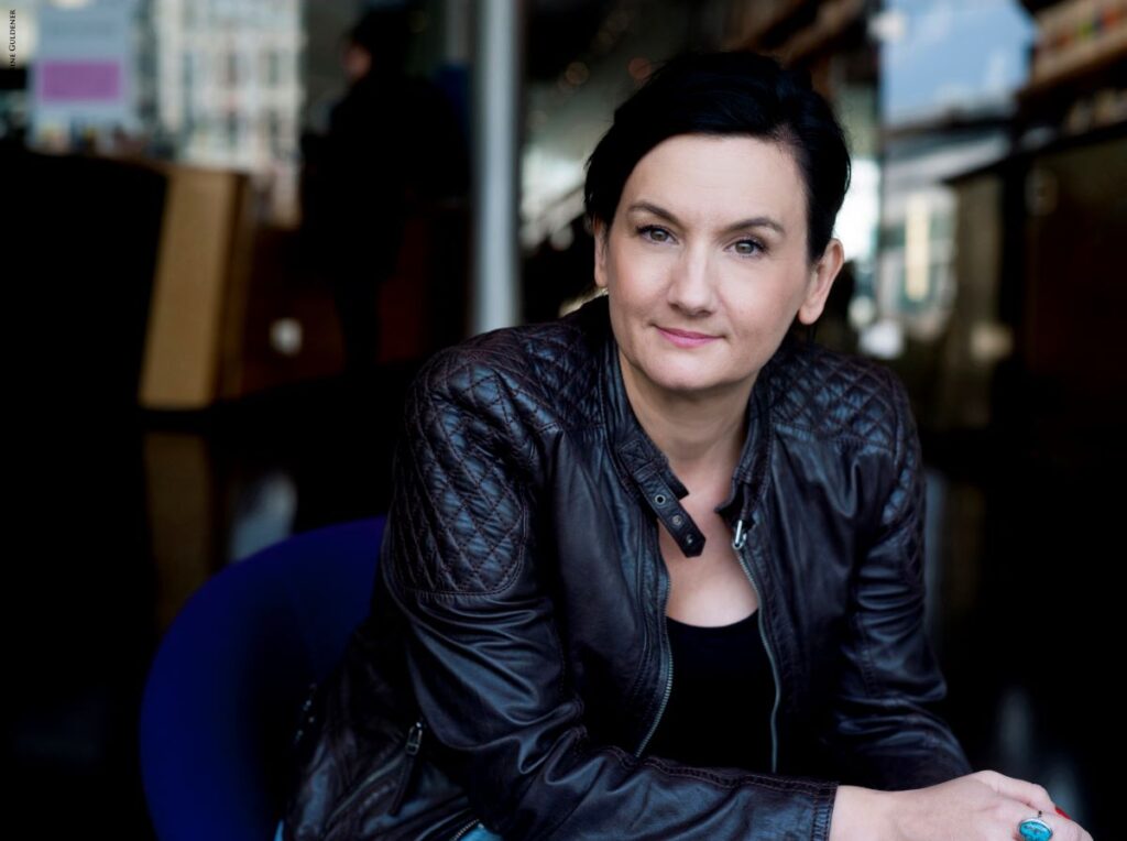 Frau mit braunem, zusammengebundenem Haar, braunen Augen und rosa Lippenstift, bekleidet mit schwarzer Lederjacke und schwarzem Shirt sitzt in einem blauen Sessel und blick in die Kamera