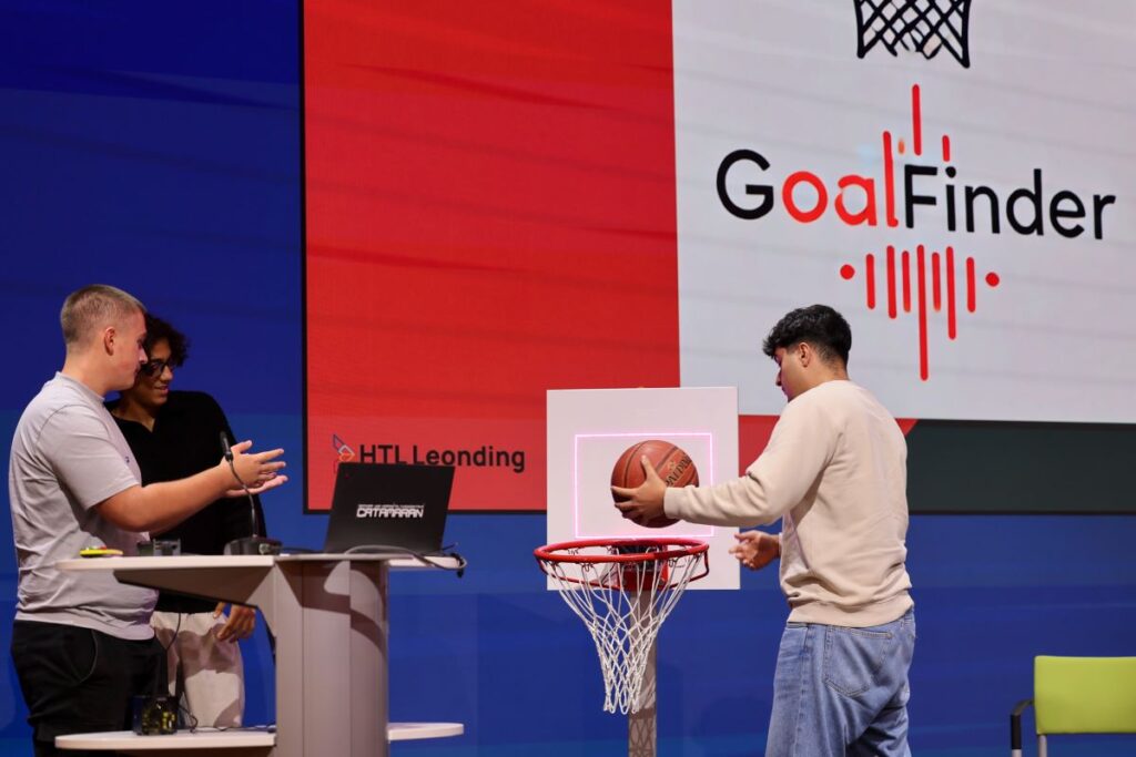 3 junge Männer präsentieren auf einer Bühne einen niedrig aufgestellten Basketballkorb