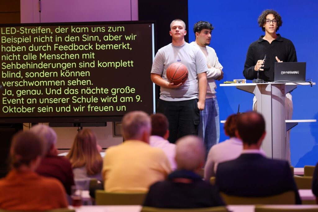 3 junge Männer präsentieren auf einer Bühne einen niedrig aufgestellten Basketballkorb. Links ist Schriftdolmetschung eingeblendet, im Vordergrund sind sitzende Personen zu sehen